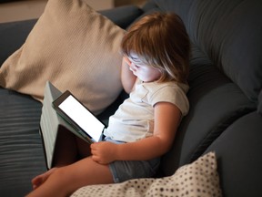 blonde three years old baby shirt and shorts, sitting comfortably in sofa inside home at night reading and watching blank screen digital tablet, face illuminated