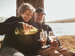 Senior couple enjoying drinks at campsite near Lake. Mature woman pouring wine in glasses, both sitting under a tree on a summer day.