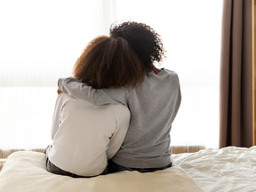 Rear back view black mother and daughter embrace sitting on bed at home, older sister consoling younger teen, girl suffers from unrequited love share secrets trustworthy person relative people concept.