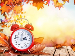 In this stock photo, an alarm clock sits on a wooden deck with autumn leaves falling.