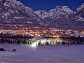 The Town of Canmore is lit up at night with fog rising from the Bow River below.