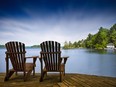 A couple of wooden Muskoka chairs sitting on the dock with a lake and cottages across in the background. Perfect for cottage related applications