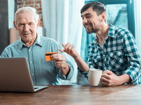 New technologies. Delighted positive young man smiling and pointing at the laptop screen while helping his grandfather to do an online payment