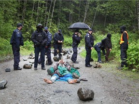 A woman plays a trumpet while lying on the road as RCMP officers assemble during an operation to arrest protesters manning the Waterfall camp blockade against old growth timber logging in the Fairy Creek area of Vancouver Island.