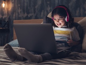 Little girl wearing headphones, sitting on bed in dark bedroom and laughing while looking at laptop.