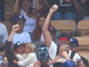 A sunglasses-wearing man with a baby strapped to his chest and beverage in hand catches a fly ball at a Los Angeles Dodgers game on April 30, 2023.