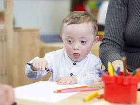 In this stock photo, a Down Syndrome child colours at a nursery with his teacher sitting next to him.