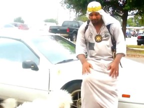 In this screenshot, Father Jesus of Gulfport, Miss. leans on a car near a parking lot.