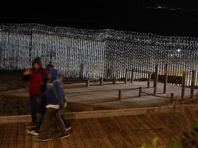 Floodlights from the U.S. illuminate the border wall, topped with razor wire, as people pass Monday, Jan. 7, 2019, at the beach in Tijuana, Mexico.