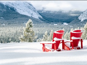 Photo of Two Snow Covered Red Adirondack Chairs Facing a Frozen Wooded Valley on a Snowy Winter Day. Banff National Park, AB, Canada.