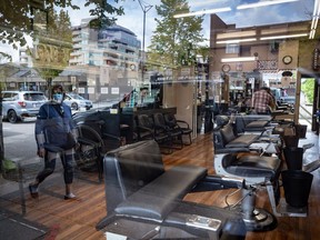 A man cleans inside a closed barbershop as a pedestrian wearing a face mask is reflected in the shop's window, in Burnaby, B.C., on Wednesday, May 6, 2020.