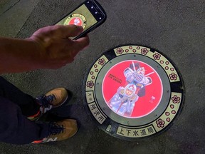 A passerby using a smartphone takes a photo of an illuminated manhole cover with designs of popular animation character Gundam, on the street in Tokorozawa, near Tokyo, Japan August 19, 2020.