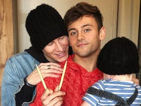 Dustin Lance Black looks on as Tom Daley holds knitting needles and son.