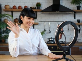 Cheerful brunette woman in stylish blouse shoots video for blog with smartphone and light ring sitting at table in kitchen