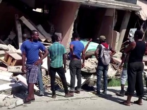 People stand in front of a collapsed building following an earthquake, in Les Cayes, Haiti, in this still image taken from a video obtained by Reuters on Aug. 14, 2021.