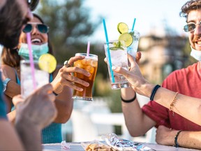 Covid-19 party. Young millennial people drinking cocktails and toasting together with a protective mask down during coronavirus pandemic at outdoor restaurant