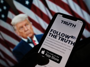 This illustration shows a person checking the app store on a smartphone for "Truth Social" with a photo of former U.S. president Donald Trump on a computer screen in the background.