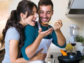 Young man getting his wife to try the food he is preparing in the kitchen at home.
