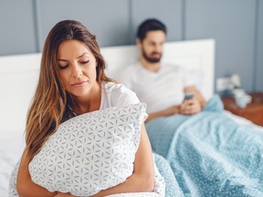 Unsatisfied woman holding pillow and sitting on the bed while her husband is on smart phone.