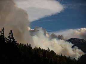 A helicopter carrying a water bucket flies past the Lytton Creek wildfire burning in the mountains near Lytton, B.C., on Sunday, Aug. 15, 2021. Two out-of-control wildfires in northeastern British Columbia that have already forced some residents to evacuate their homes are expected to grow bigger in the next few days.