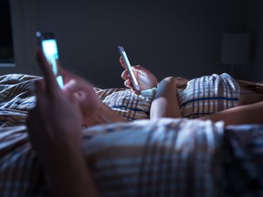 Bored, distant couple ignoring each other while lying in bed at night using their phones.