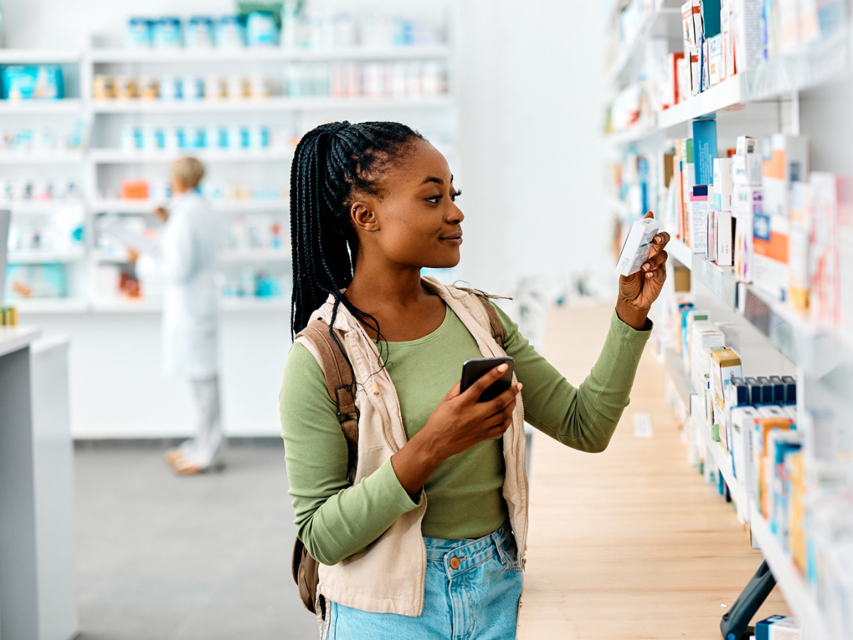 Young African American woman using cell phone while looking for a medicine in drugstore.