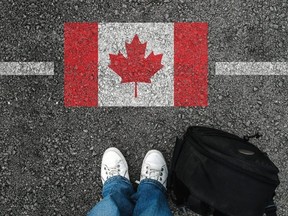 A man with a shoes and backpack is standing on asphalt next to flag of Canada and border.