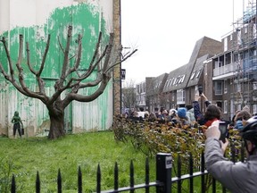People look at a new Banksy painting on a wall in London, Monday, March 18, 2024. A new Banksy mural drew crowds to a London street on Monday, even before the elusive graffiti artist confirmed that the work was his. The artwork in the Finsbury Park neighborhood covers the wall of a four-story building and shows a small figure holding a pressure hose beside a real tree. Green paint has been sprayed across the wall, replicating the absent leaves of the tree, which has been severely cropped. Banksy claimed the work by posting before and after photos of the location on his official Instagram account.