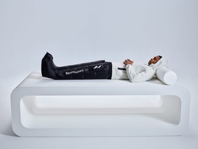 Adult laying horizontally on an elevated platform, wearing a white sweatshirt and the black Normatec compression leg boots. They extend from the toes to the hips.