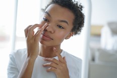 Attractive afro girl is applying cream touching her face. Femininity and grooming concept. Mirror reflection of young latin woman. Skin cleansing. Daily routine, cosmetics and hygiene of teenage girl.