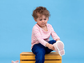 A child sitting on a yellow crate and putting on a pair of pink rainboots.