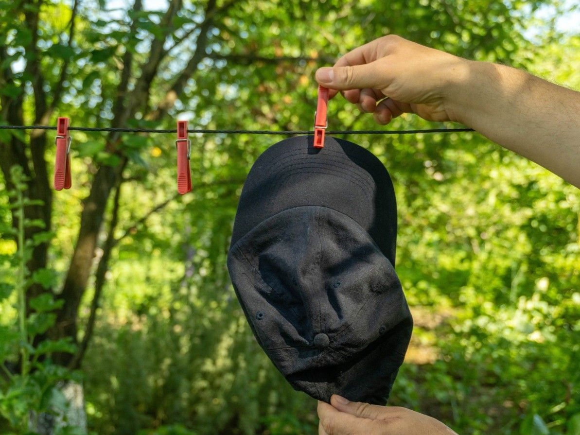 A hand clips a black hat onto a clothesline using red clothespins. The background is filled with green plants and trees under a clear sky during the day