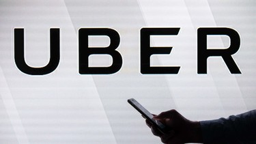 A man checks his smartphone while standing against an illuminated screen bearing the Uber logo in London on June 26, 2018.