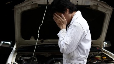Frustrated stressed young mechanic man in white uniform covering face with hands with car in open hood at the garage.