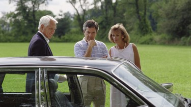 A white-haired gentleman trying to sell an older car to a couple.
