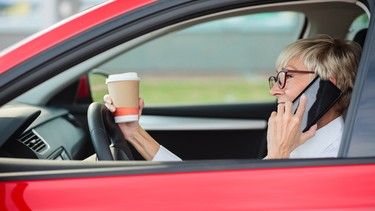 Reckless, smiling mature woman talking on the phone and holding a cup of coffee while driving a car