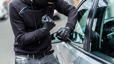 A man dressed in black with a balaclava on his head trying to break into a car.