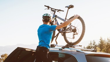 A mountain biker lifting a bike onto a roof-mounted rack on a Mercedes-Benz SUV
