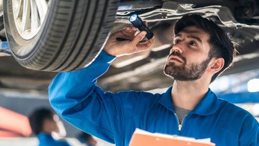 handsome man mechanic checking under car condition in garage. Automotive mechanic pointing flash light on wheel following maintenance checklist document. Car repair service concept