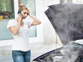 Young woman using mobile phone while looking at broken down car on street breakdown coolant steam smoke