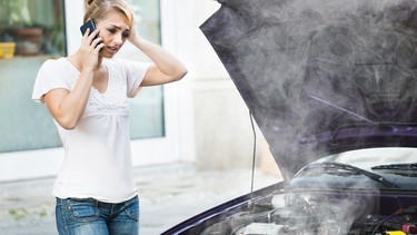 Young woman using mobile phone while looking at broken down car on street breakdown coolant steam smoke