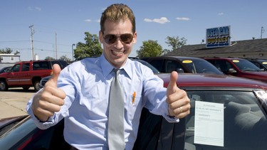 A salesman giving a thumbs-up in a used-car dealership parking lot.
