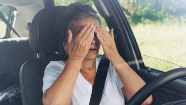 Mature woman driver sitting at the wheel in a car, frightened of a road accident and covering her eyes with her hands, view through the windshield. Road traffic accident concept