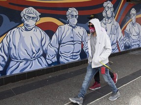 A man wearing a protective face mask walks past a mural of heath care workers painted on a boarded up restaurant in downtown Vancouver, Monday, April 27, 2020.