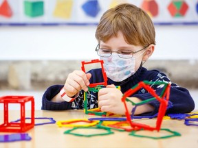 Little child with medical mask playing with lots of colorful plastic blocks kit in preschool nursery or elementary school. Cute child use protective equipment as fight against covid 19 corona virus.