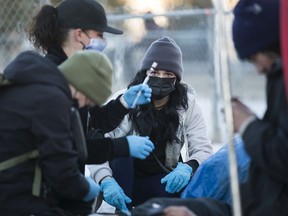 A person holds a syringe while two others, all wearing winter gear and laytex gloves, assist a person laying on a sidewalk