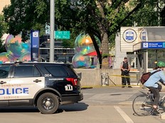 A police officer stands on a sidewalk with crime scene tape visible. A police vehicle is in the foreground and a cyclist rides past the scene.