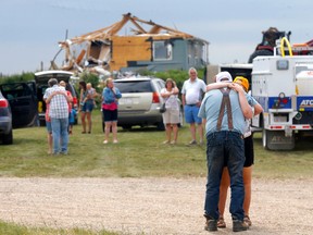Two people hug as a crowd of people gather by a destroyed farm structure in the background