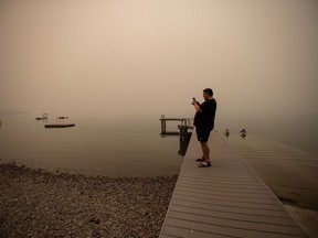File photo: Thick smoke from wildfires blankets the area as a man standing on a pier takes a photo of Okanagan Lake, in Lake Country, B.C., Friday, Aug. 13, 2021. Evacuation alerts and a state of local emergency caused by a wildfire on Knox Mountain in Kelowna have ended.
