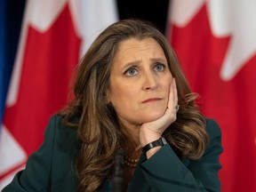 Chrystia Freeland sits at a podium with chin in hand, Canadian flags in the background